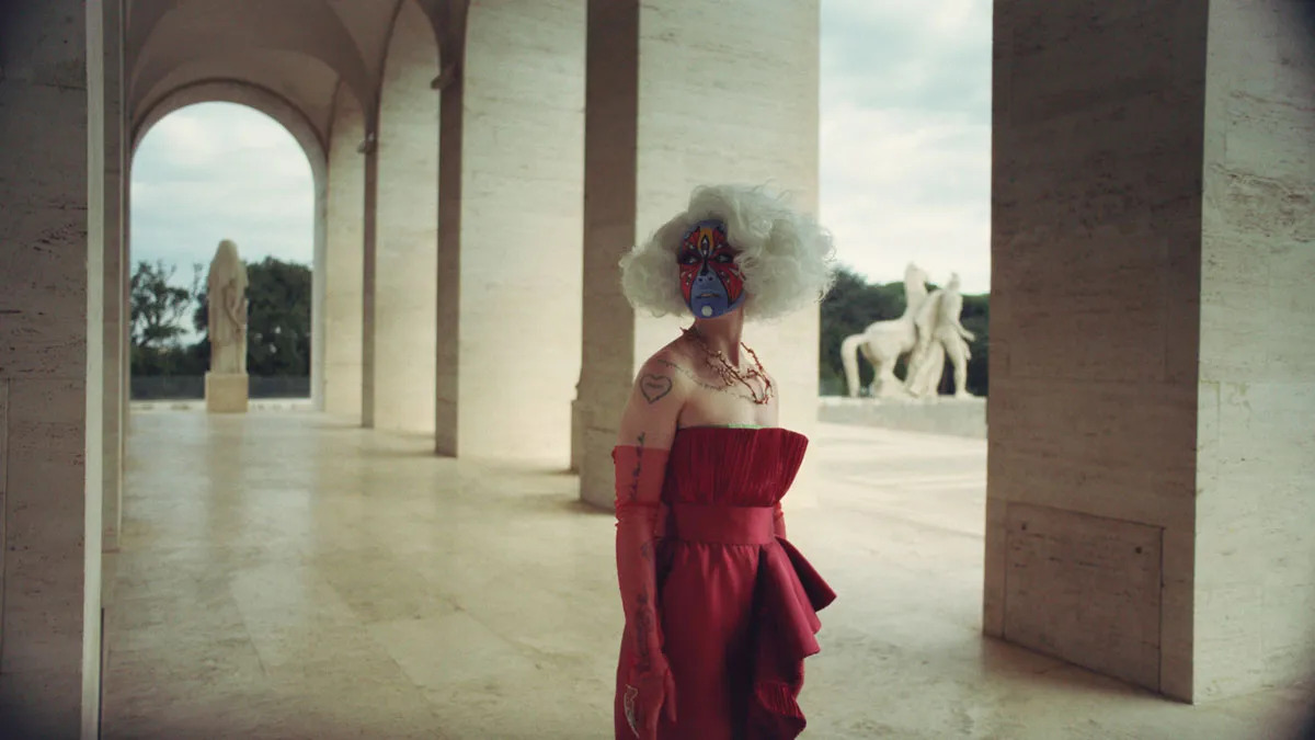 A woman in a red dress with a white wig and blue-and-red face makeup walks through a marble outdoor hallway.