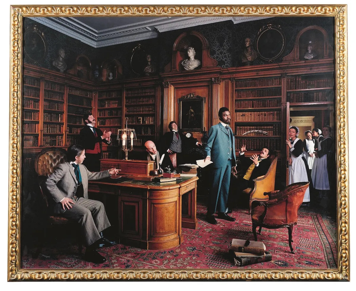 A photograph of a Black man standing amid white men and women in an old-fashioned library.