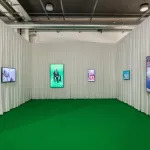 A booth whose walls are covered in white curtains, to which five screens have been hung. The floor has been painted green.