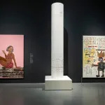 Installation view of a museum exhibition showing two paintings on the wall with a sculpture of a white column that is carved into in the center.