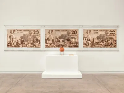 Three giant bank notes hang on a gallery wall behind a terracotta vase.