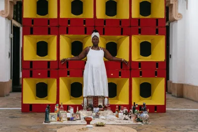 A Black woman in a white dress stands in front of a home altar on the floor.