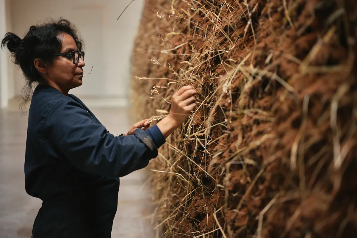 Delcy Morelos, wearing a blue jumpsuit, places hay into a large sculpture made of earth.