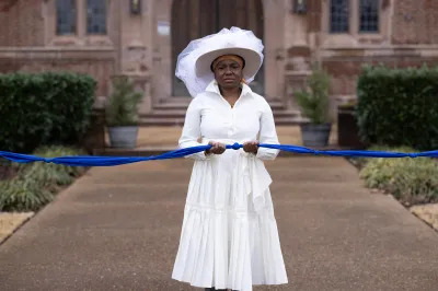 A Black woman in all white holds a long rope of knotted blue cloth in front of a brick building. 