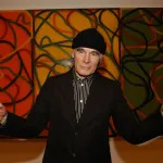 A man in a black beanie and suit jacket with his arms outstretched. He stands before a large abstraction.