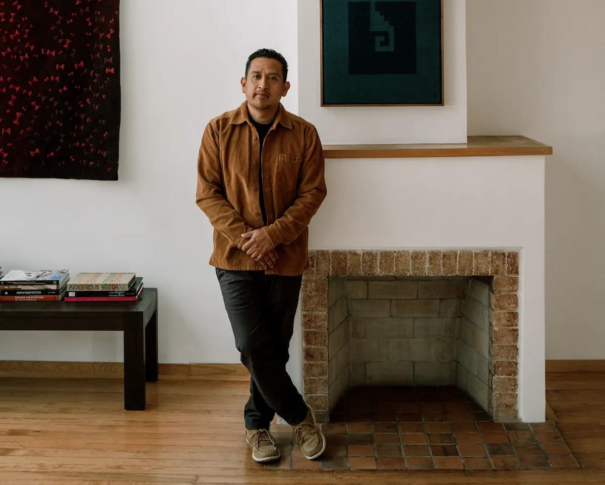 A Brown man wearing a brown shirt and pants standing with his arms held at his waist beside a fireplace. Beside him is a bench with books stacked on it, and behind him are abstract paintings on the walls.