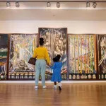 A Black mother and daughter stand before a five-panel artwork.