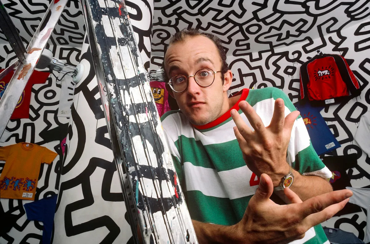 A white man in a striped green shirt holds his hands and makes a confused face. He is in a room that is lined with intersecting figures in black silhouettes; it has a pole in its centered that has been graffitied with black. Hanging on its walls are vibrantly colored T-shirts.