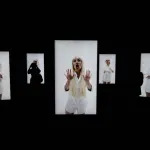 Silhouetted figures stand in front of backlit boxes displaying images of a white woman with a blonde wig. In one, she presses her hands together; in another, she holds her hands up.