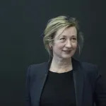 Portrait of a white woman with blonde-ish hair in a navy blazer and black blouse on a green-ish background