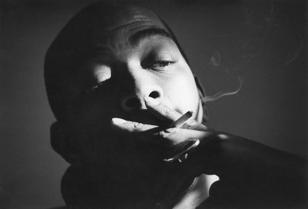 A dramatically lit black-and-white photograph of Geoffrey Holder smoking.