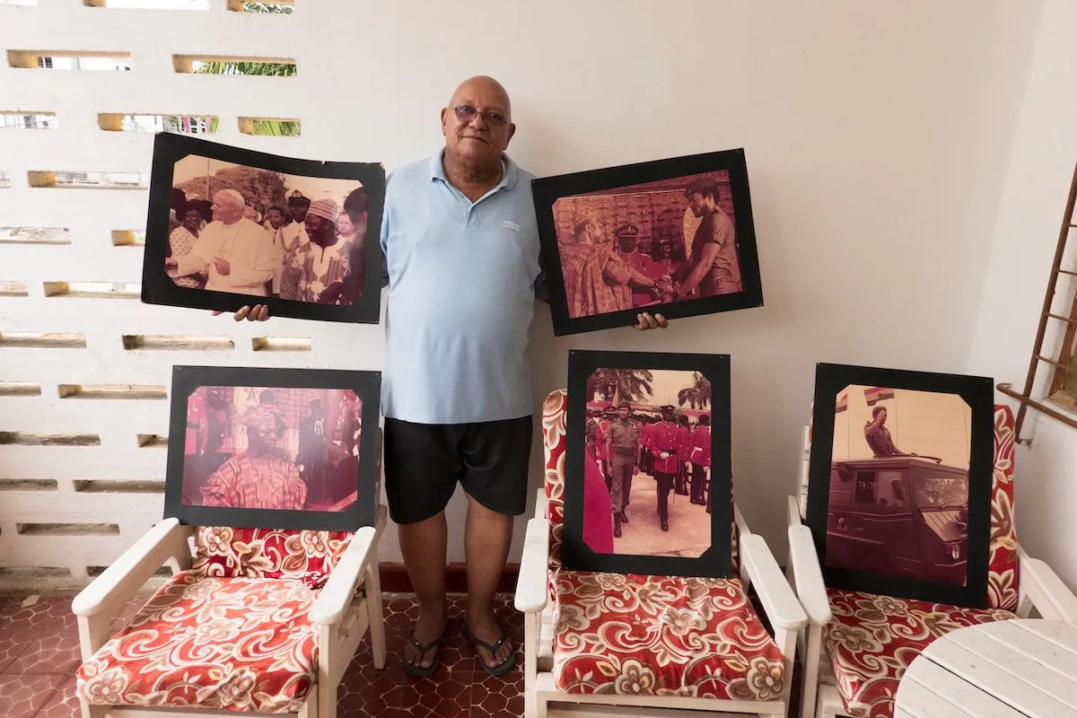 A Black man holding photographic prints, some of which are propped on chairs.