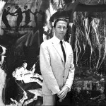 In a black-and-white photo, Robert Rauschenberg in a suit and dye stands in front of a dense painting.