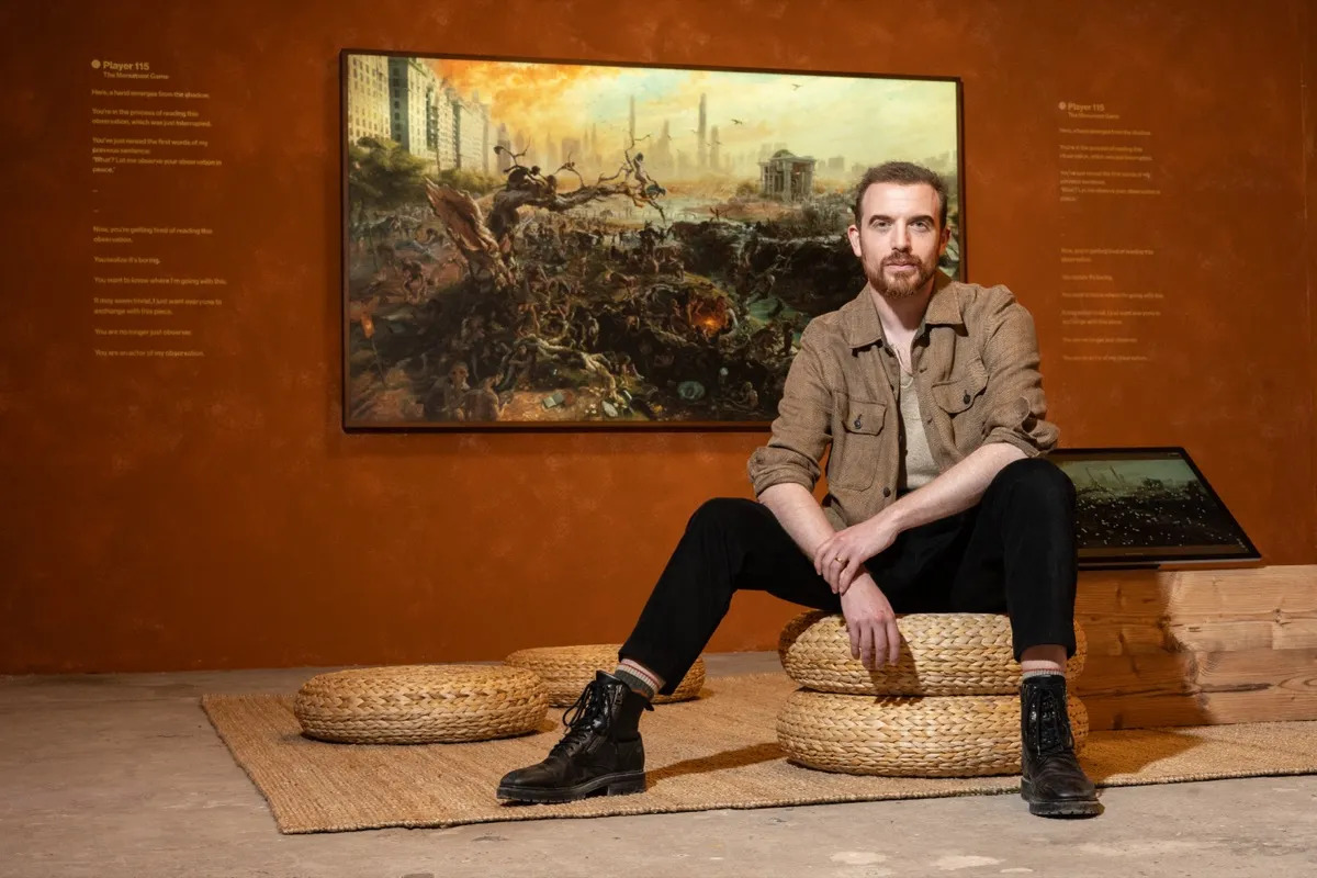 A white man with a goatee and short hair sits in front of a digital painting.