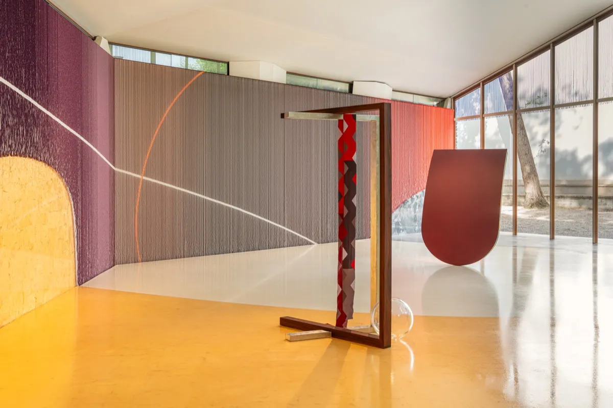 An unduluating colorful shape of beads sits behind two sculptures on a yellow floor.