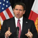 A white man holding up his two pointer fingers and standing before an American flag at a podium with a microphone.