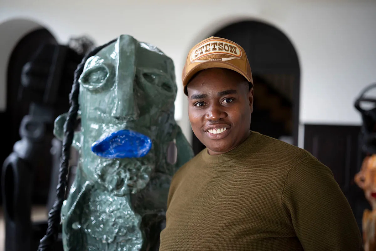 A Black woman in a baseball cap standing beside a clay sculpture with gaping blue lips.