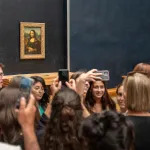 A crowd of people, some holding smartphones, stands before a painting of a woman with her arms in her lap.