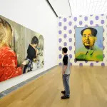 A man staring at a long painting of two women. On an adjacent wall, an image of Chairman Mao hangs above purple polka dots.