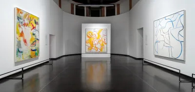 View of a museum exhibition showing three abstract paintings on hanging on different walls. 