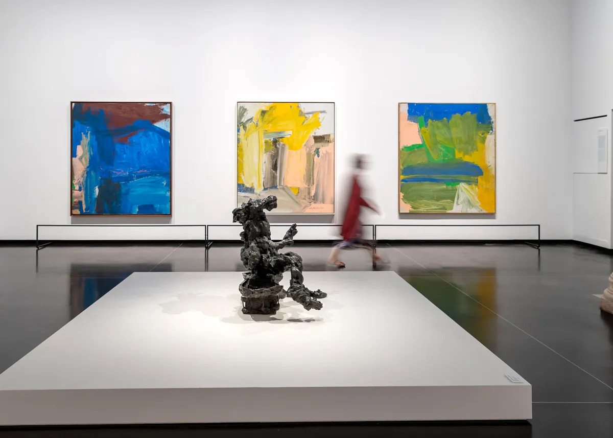 View of a museum exhibition with three abstract paintings on the same size hanging on a wall and a bronze sculpture on a low plinth in the center of the room. A blurred figure walks between them.