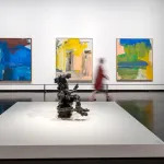 View of a museum exhibition with three abstract paintings on the same size hanging on a wall and a bronze sculpture on a low plinth in the center of the room. A blurred figure walks between them.