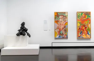 View of a museum exhibition with a bronze sculpture on a two tier plinth in a corner and two semi-figurative paintings to the right on a wall. 