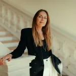 A woman with brown hair in black blazer leans against a white bannister in front of stairs.