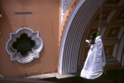 A Latina woman in a shroud being hoisted by a harness before an arch.