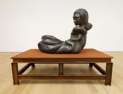 A sculpture of a twisted black human form exhibited atop a table-lake structure.