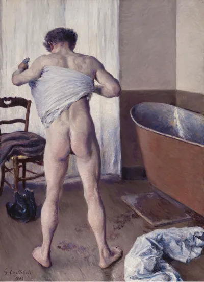 A nude white man seen from behind, wiping his back using a towel near a bath.