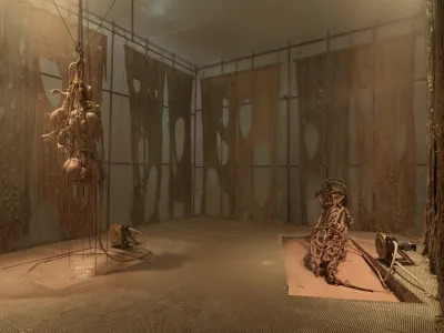 A fog-filled installation whose gridded walls are draped with material resembling torn fabric. On the floor and hanging from the ceiling are sculptures resembling guts that are attached to motors.