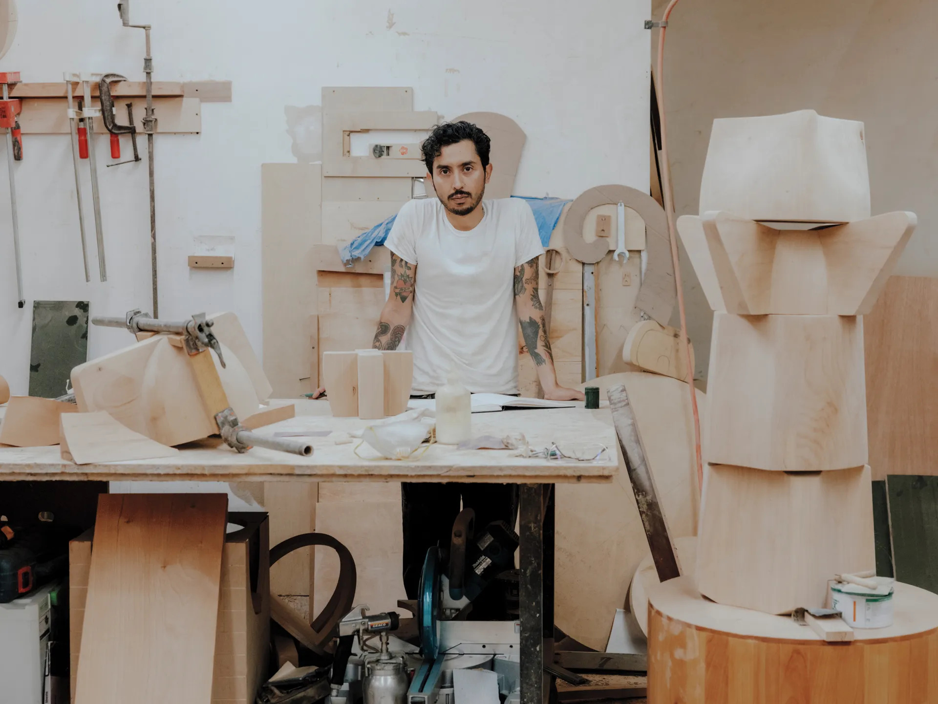 A portrait of Ryan Preciado, standing behind a table, surrounded by various sculptures in wood.