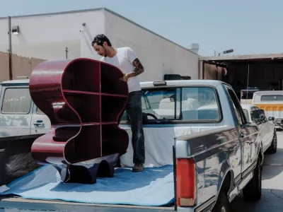 A man stands on the bed of a blue pickup truck as he secures a dark red curved sculpture. 