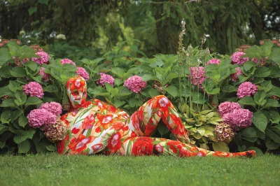 A person whose body is completely wrapped in a floral fabric lies in a grassy area surrounded by bushes and pink flowers.