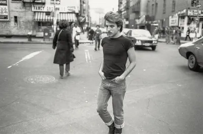 A black-and-white photograph of a mustached man in tight jeans walking on a city street.