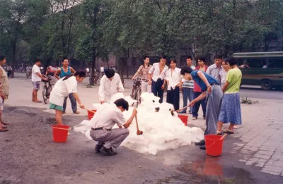 A group of people, most of whom are women, hacking at a pile of snow with shovels and pails.