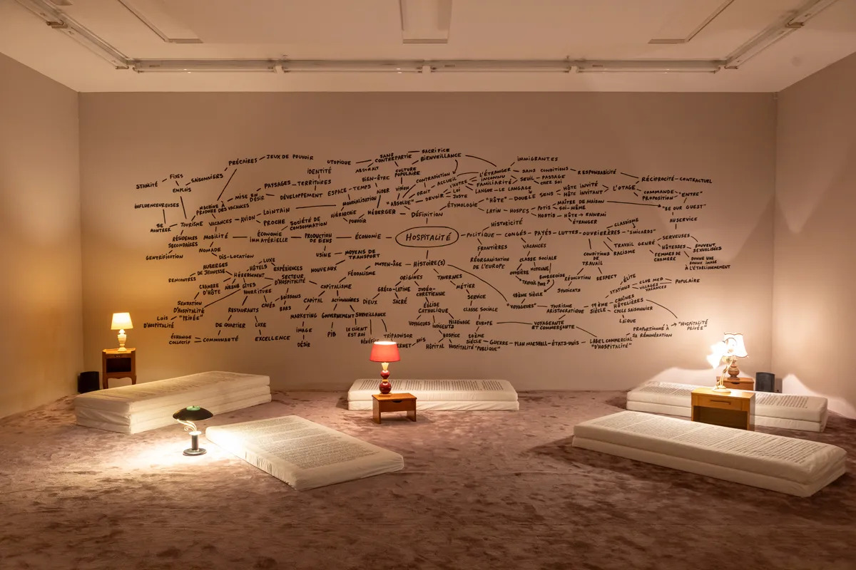 View of an installation with a word map on one wall and a series of mattresses on the floor with lamps.