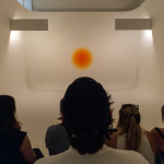 A crowd looks on at a yellow-org circle in a white cylindrical background