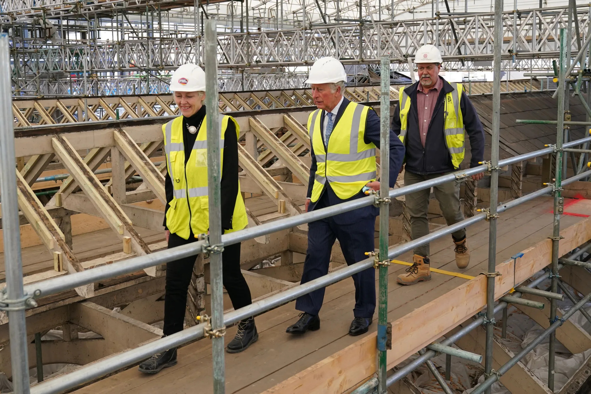 Three people in high vis jackets and hard hats walk on scaffolding.