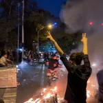 A woman holds up her hands in two Vs looking at a fire during a protest on a busy street in Tehran.