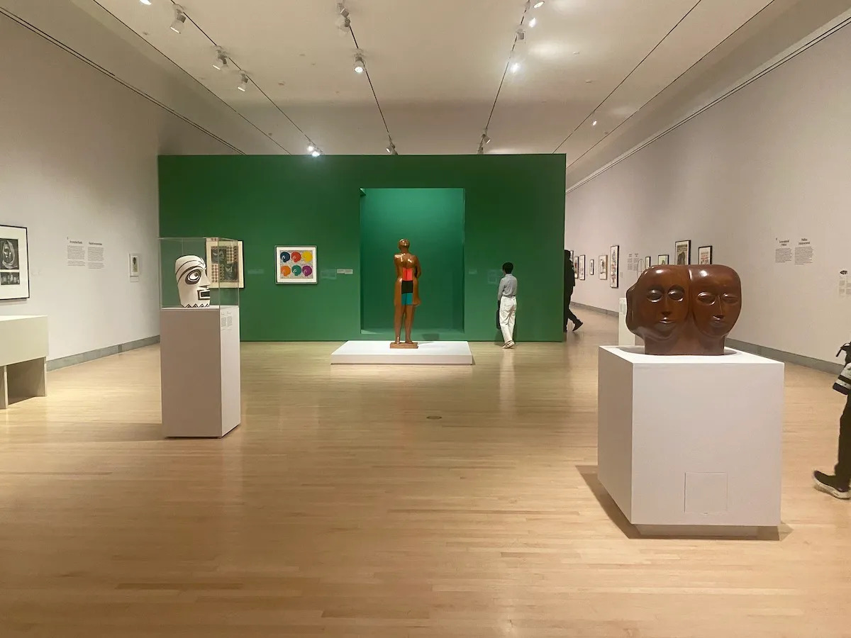 A museum gallery with a prominent green wall at its back and, at its center, a sculpture on a pedestal that has two heads put side by side.