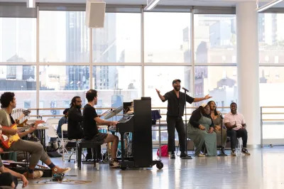 A man sings standing at a mic, with three backup singers seated at right and at left a pianist, dummer, and other musicians. 