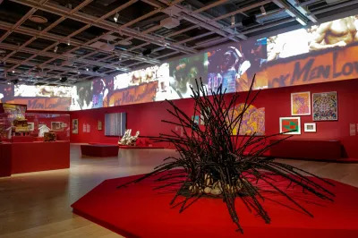 View of a museum exhibition with red walls and screens at the top. A sculpture of wood is seen in the foreground. 