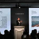 A man stands on a rostrum during an auction in front of two digital screens.