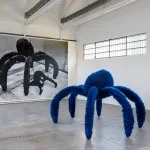 A sculpture of a fuzzy blue spider with a black-and-white picture of a man pretending to do a somersault beneath the sculpture.