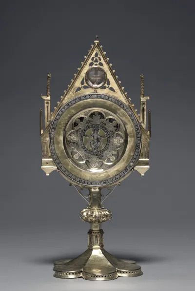 Monstrance with the "Paten of Saint Bernward", circa 1180-1190. Designed to facilitate the display and veneration of ten relics, this sumptuous ostensorium (from the Latin ostendere, meaning "to show") is one of the most unusual reliquaries in the Guelph Treasure. Elements of contemporary Gothic architecture in miniature frame an elaborately decorated liturgical paten, the shallow disc or plate used for the elevation of the Eucharist during Mass. Fragments of the True Cross are enclosed behind a rock crystal in the gable, while other saintly relics, each wrapped in silk and identified by inscriptions, are visible only from the back. The paten is inscribed on its outer rim with the words "The bread which is broken in me is the body itself. He who receives it in good faith shall live in eternity." Another inscription surrounding the image of the enthroned Christ at the paten