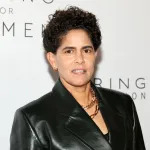 A woman poses in front of a white wall, wearing a black leather jacket. and necklaces.