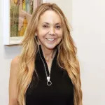 Portrait of a smiling white woman in a black top with a long zipper running down its middle. Behind her are abstract paintings.