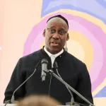 A Black man dressed in black stands on a podium with a microphone.